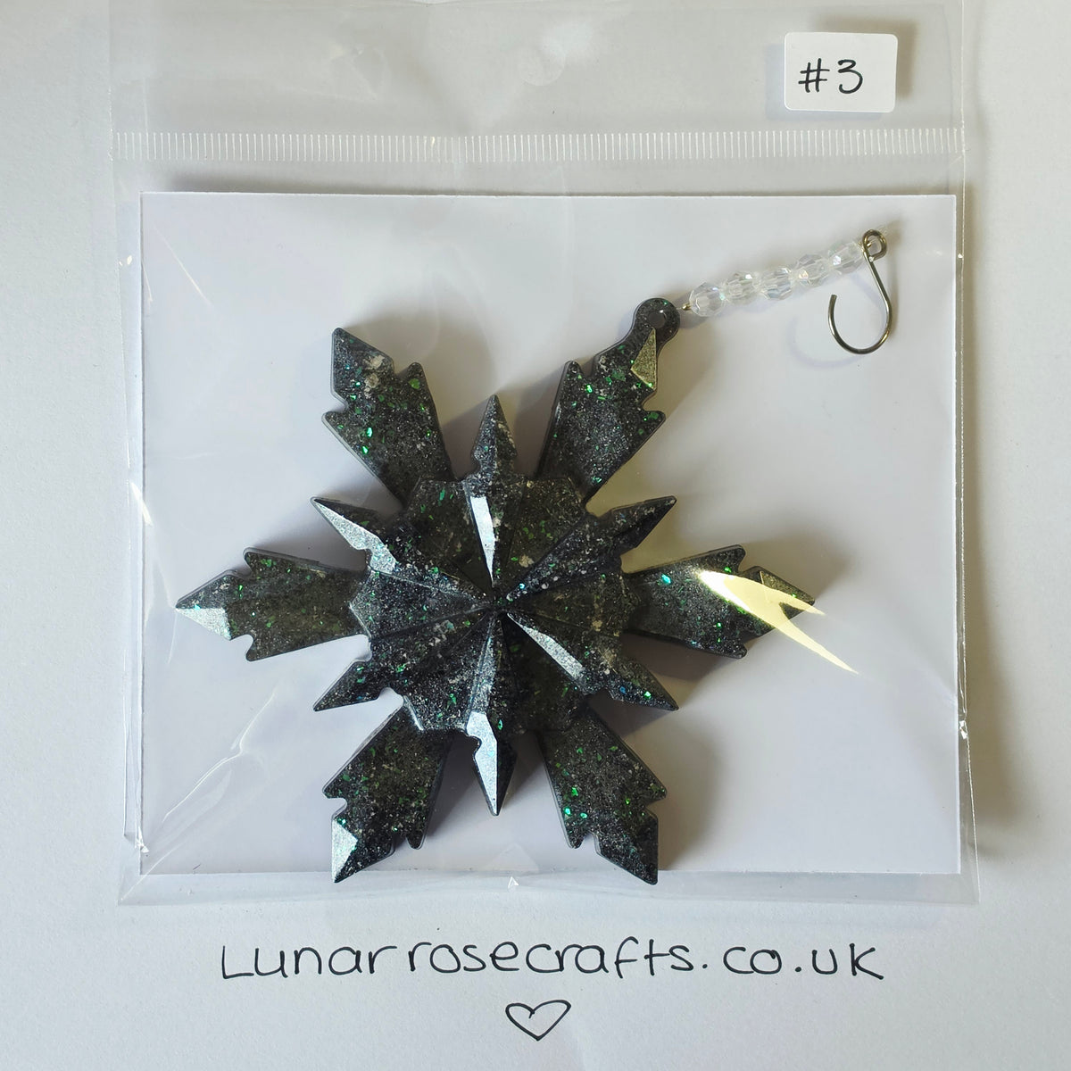 A glittery black snowflake ornament with silver glitter accents and a clear bead loop, packaged in a clear plastic bag on a white backing card. The card has the Lunarrosecrafts.co.uk