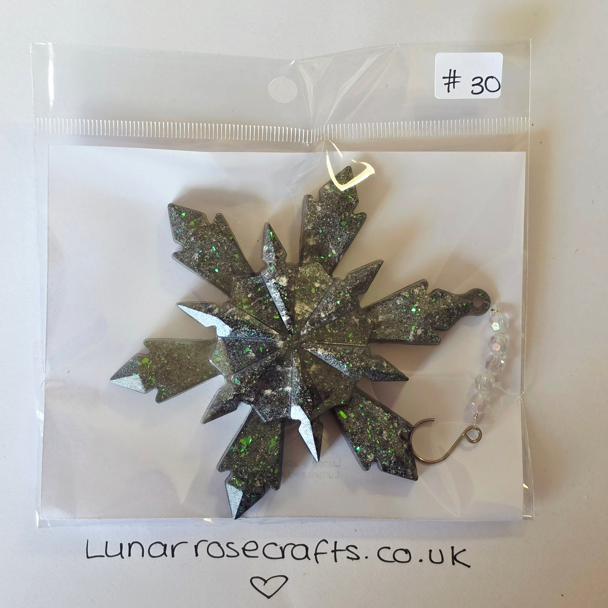 A glittery snowflake-shaped hanging decoration oin a clear plastic bag with a handwritten price tag '#30' and the website 'Lunarrosecrafts.co.uk'