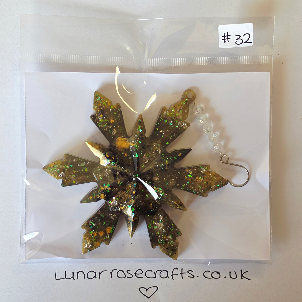 A glittery star-shaped ornament in olive green and gold, sealed inside a clear plastic bag with a white backing card. A small handwritten label reads '#32' and the card shows the brand name 'Lunarrosecrafts.co.uk'