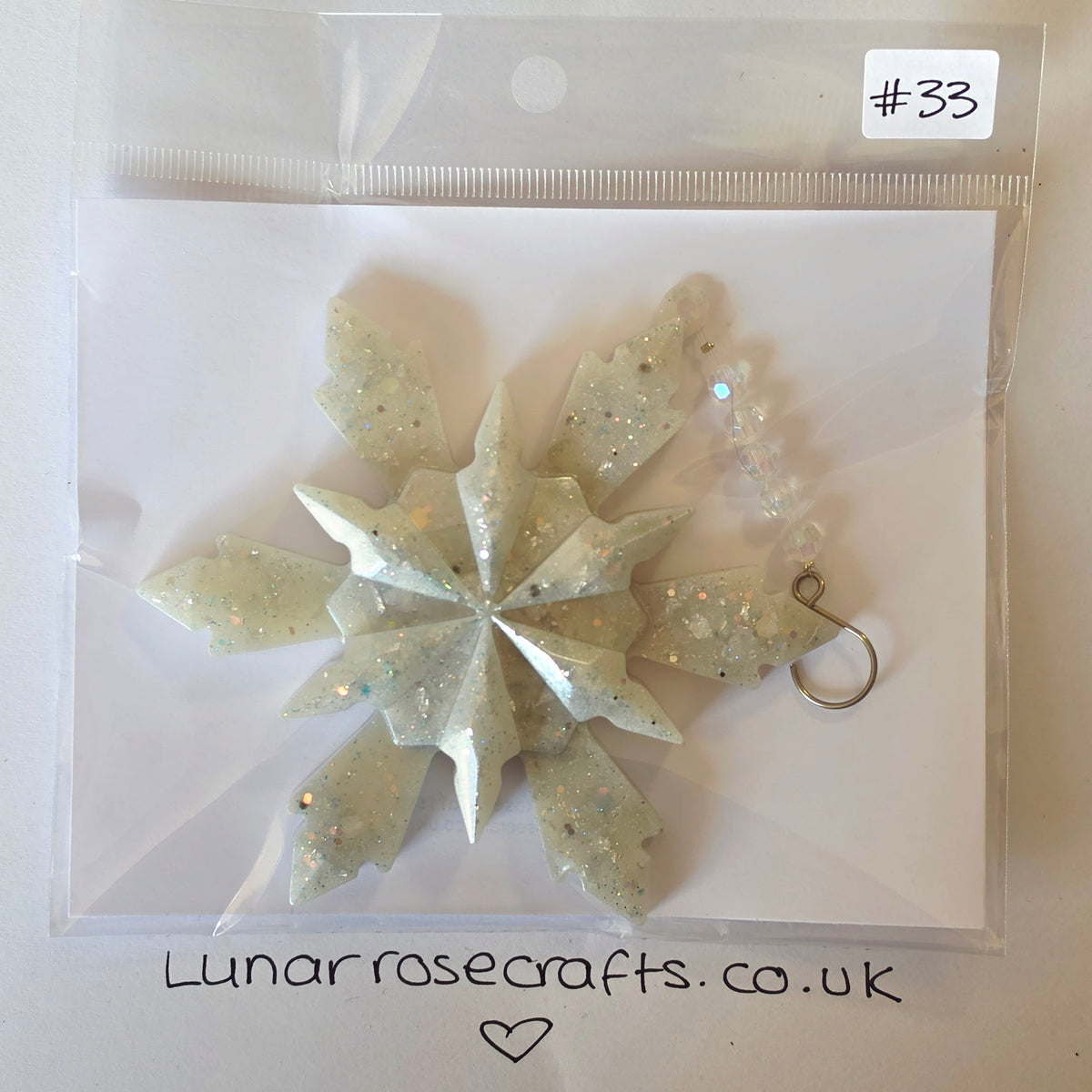A packaged white glittery snowflake ornament with a small metal loop, sealed in a clear plastic bag on a white backing card. A handwritten label reads #33, and Lunarrosecrafts.co.uk