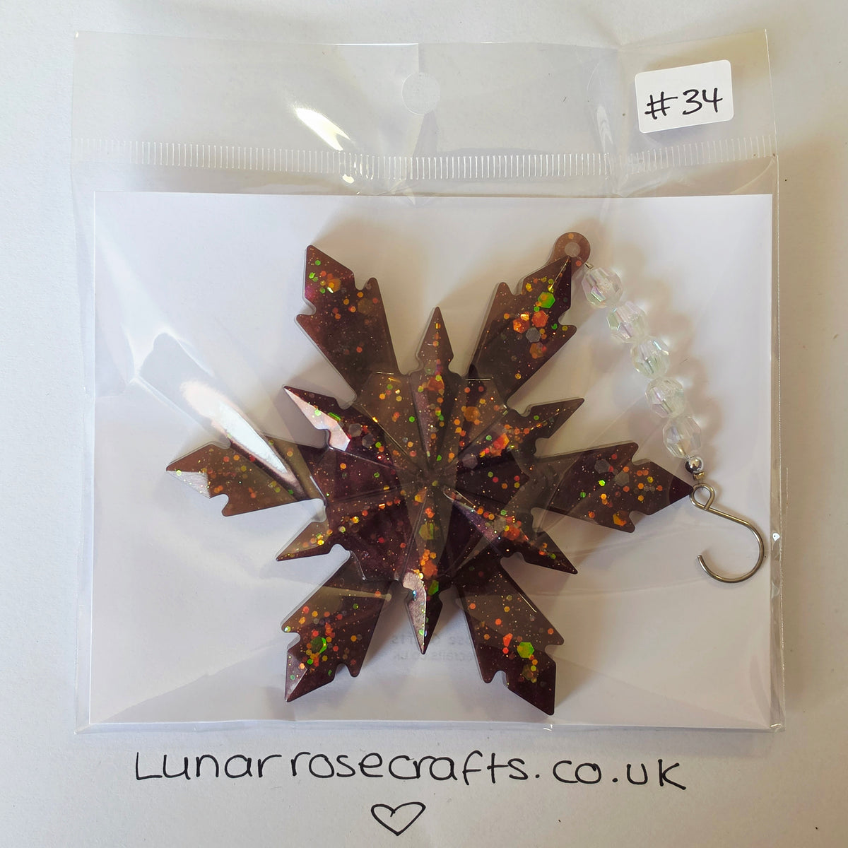 A glittery snowflake-shaped hanging decoration in dark brown resin with orange, green, and red flecks, packaged in a clear plastic bag on a white backing card. A small handwritten label says '#34' and the card is branded 'Lunarrosecrafts.co.uk'