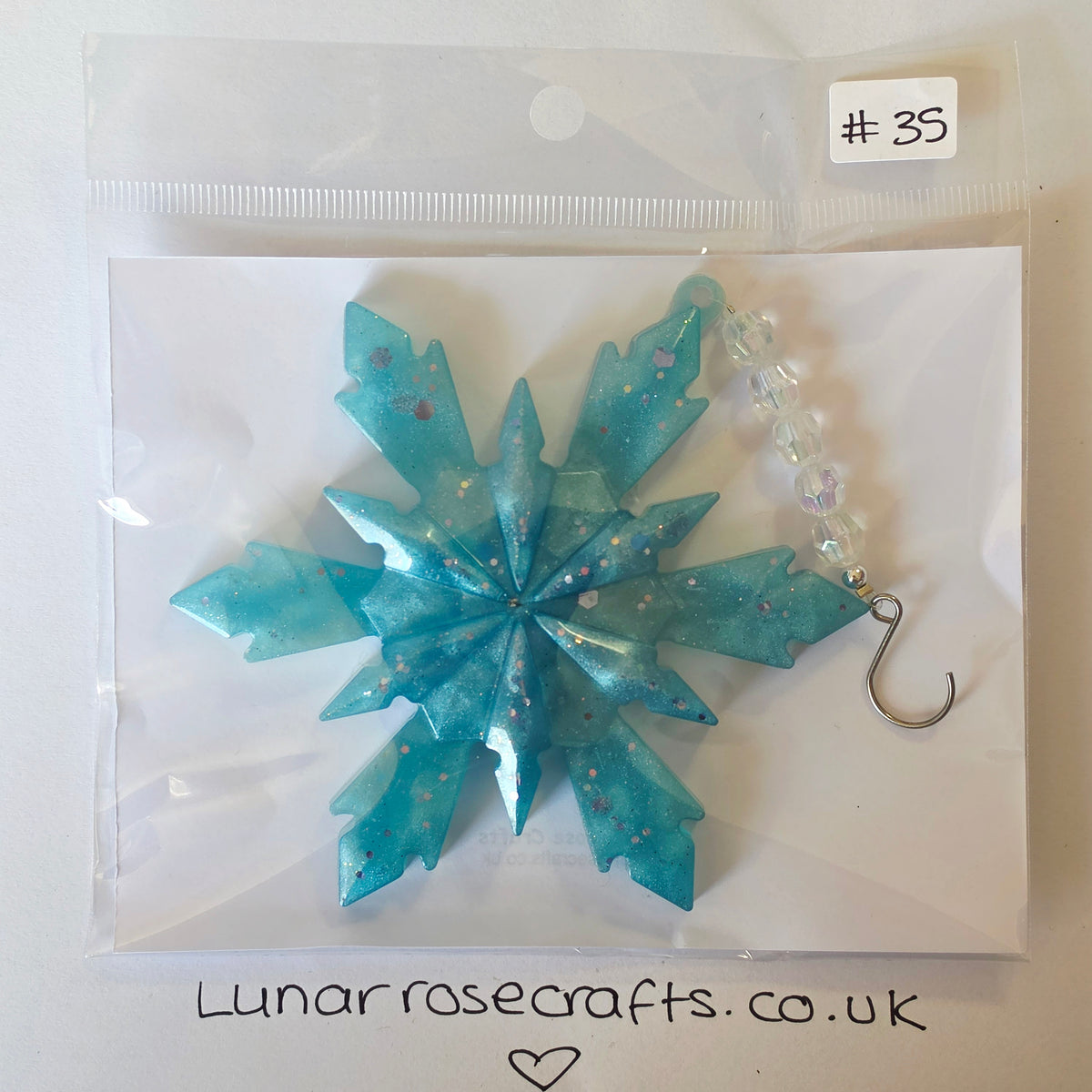 A handmade snowflake-shaped hanging decorationin translucent aqua blue resin with silver glitter, packaged in a clear plastic bag with a white backing card. A silver hook charm with clear faceted beads is attached.