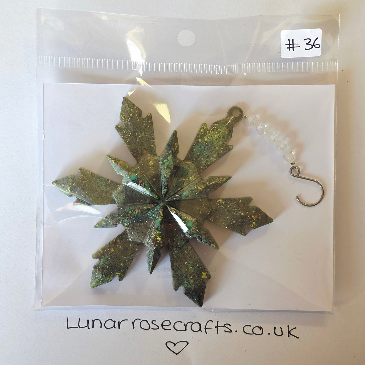 A glittery snowflake-shaped hanging decoration in olive green and gold, packaged in a clear plastic bag with a white backing card. A small white label reads '#36' and the card has 'Lunarrosecrafts.co.uk'