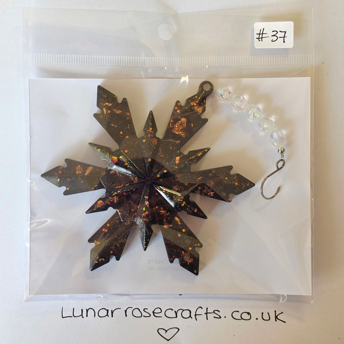 A snowflake-shaped resin hanging decoration with a dark metallic bronze finish and orange glitter flecks, packaged in a clear zip-top bag with a white card insert. A beaded hanging loop is attached, and a handwritten label '#-37' is visible on the bag.