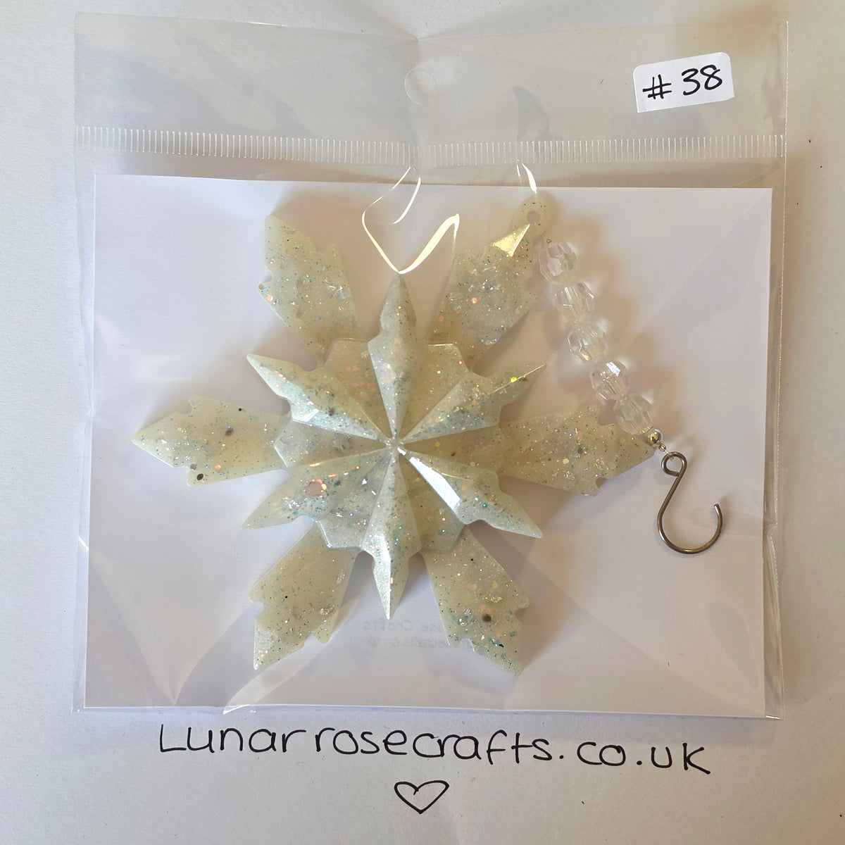 A handmade snowflake-shaped resin ornament with embedded glitter, packaged in a clear plastic bag on a white backing card. The ornament has a silver-tone metal hook.