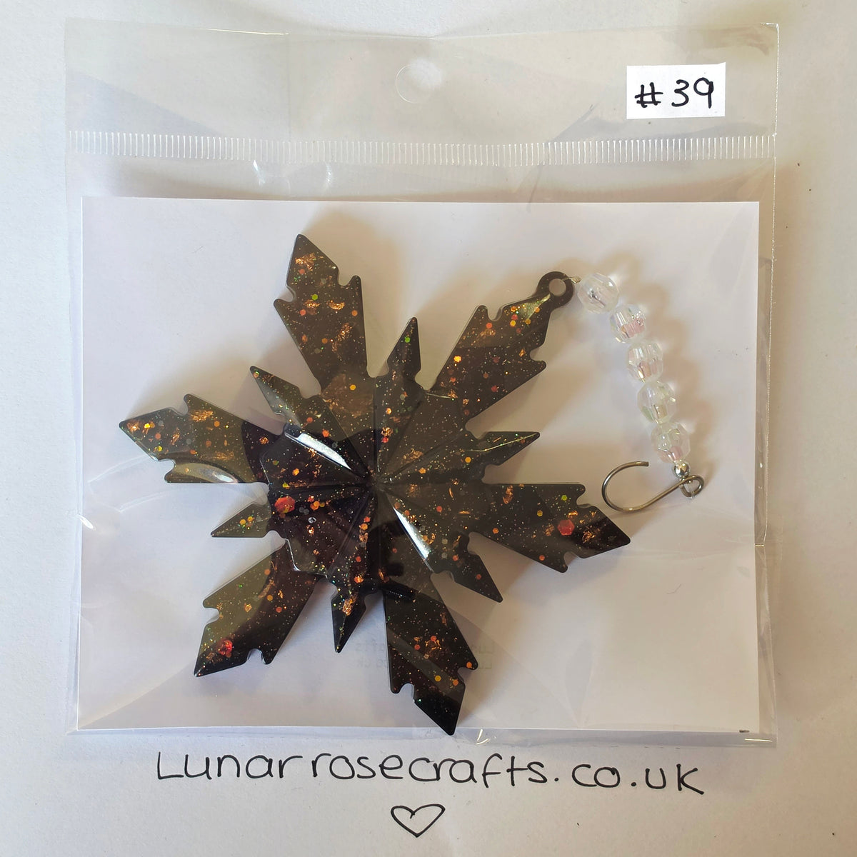 A snowflake-shaped dark brown resin hangie with orange and gold glitter flecks, packaged on a white card inside a clear plastic bag with a handwritten label '#39' and 'Lunarrosecrafts.co.uk'
