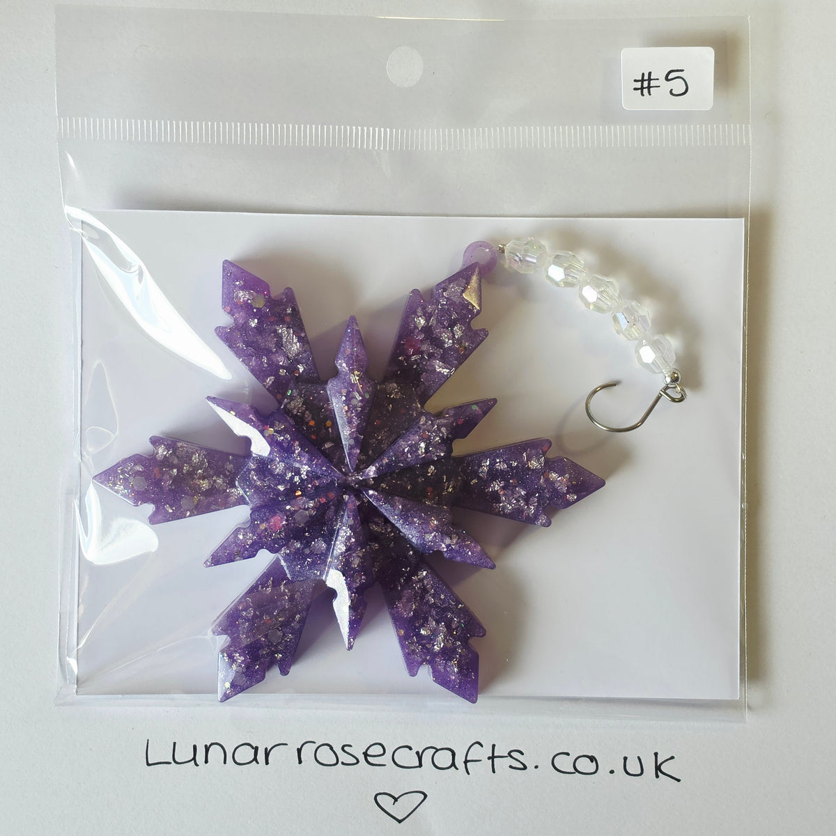 A purple glittery snowflake shaped hanging decoration displayed inside a clear plastic bag with a white backing card. A white bead chain with a silver hook is attached, and a small label reads '#5'.