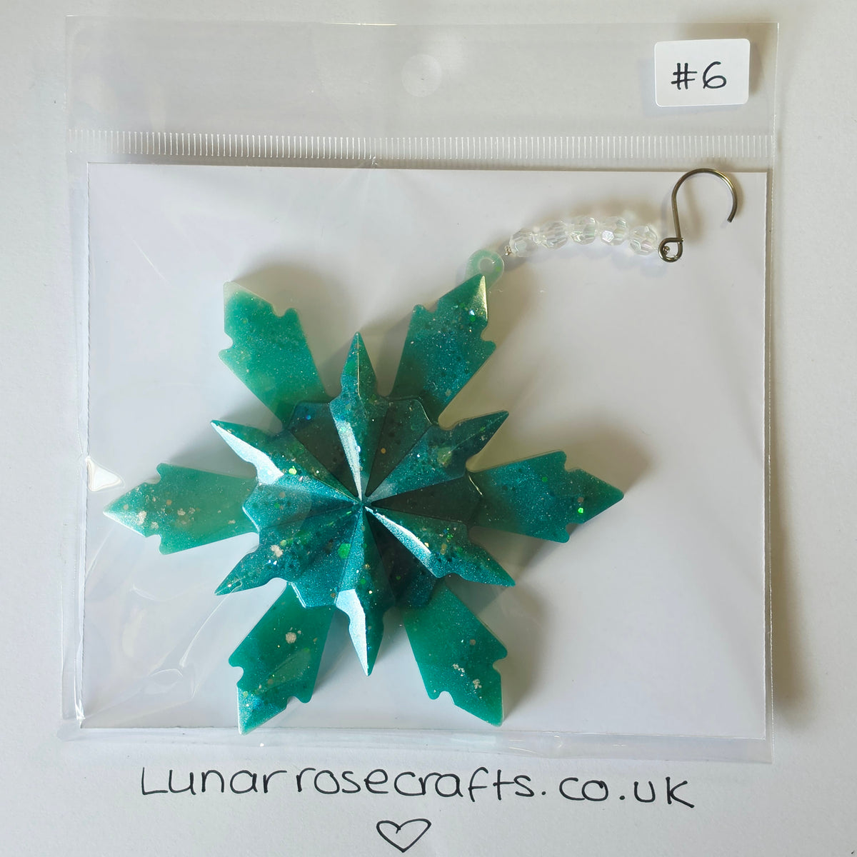 A teal green glittery snowflake ornament in a clear plastic bag with a white backing card. The bag has a handwritten label '#6' at the top right and 'Lunarrosecrafts.co.uk'