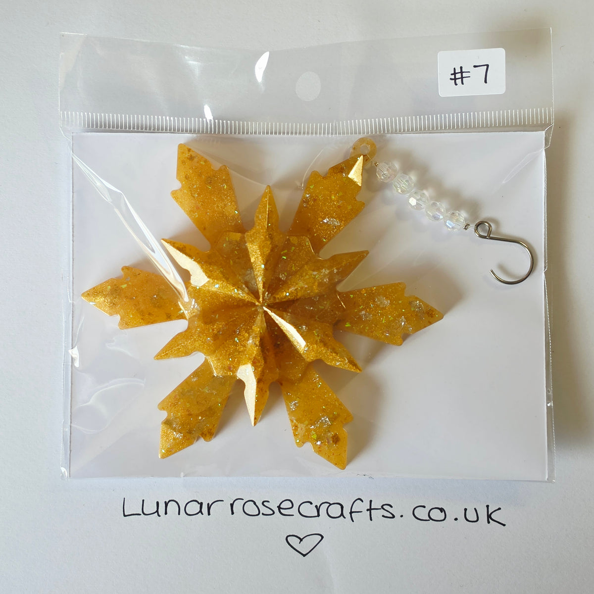 A glittery gold snowflake hanging decoration inside a clear plastic zip bag on a white backing card, with a small white label reading '#7' and a handwritten website at the bottom.
