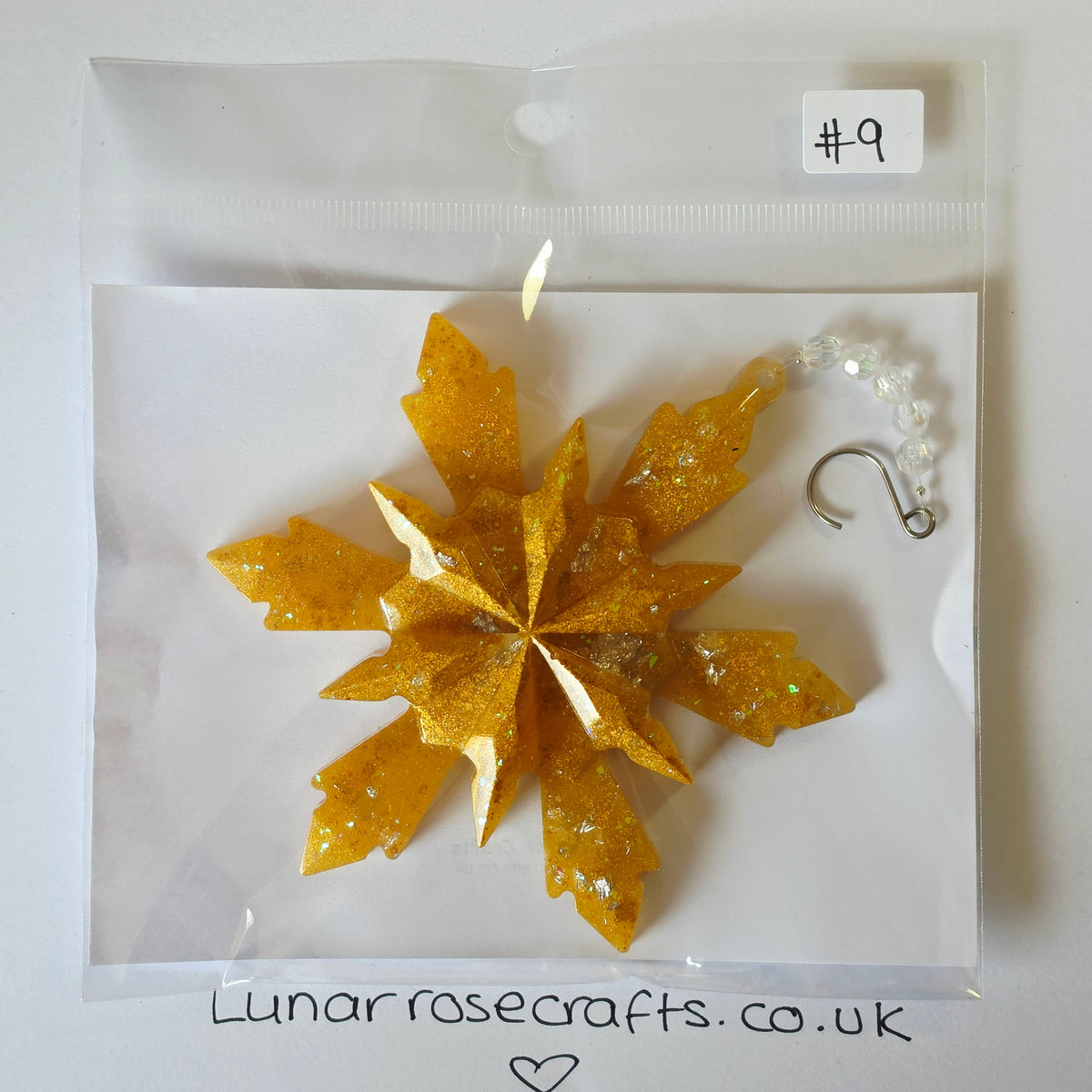 A glittery gold snowflake hanging decoration in a clear plastic bag with a white backing card. A small handwritten label says '#9' and the card shows the brand name 'Lunarroscrafts.co.uk'