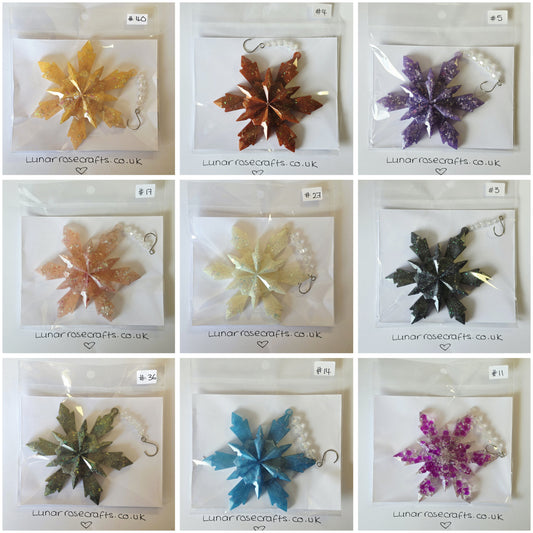 A 3x3 grid showing nine different glittery  snowflake star-shaped hanging decorations in various colours, each individually packaged in a clear plastic bag with a white backing card. The cards are labeled with product codes and the brand name 'Lunarrosecraftsco.uk