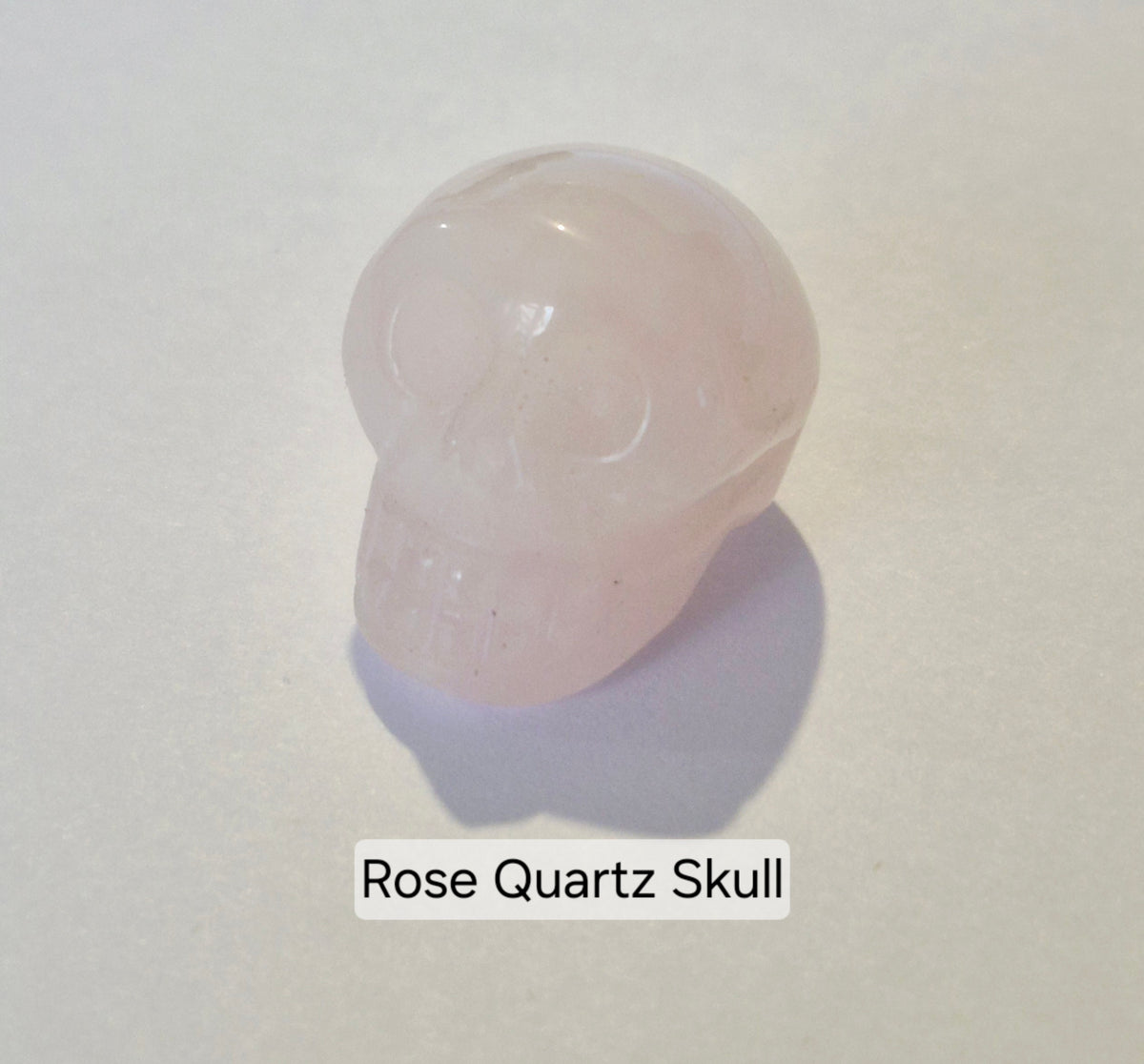 A carved rose quartz skull figurine on a light off-white background with a soft shadow below it and a text label reading 'Rose Quartz Skull'.