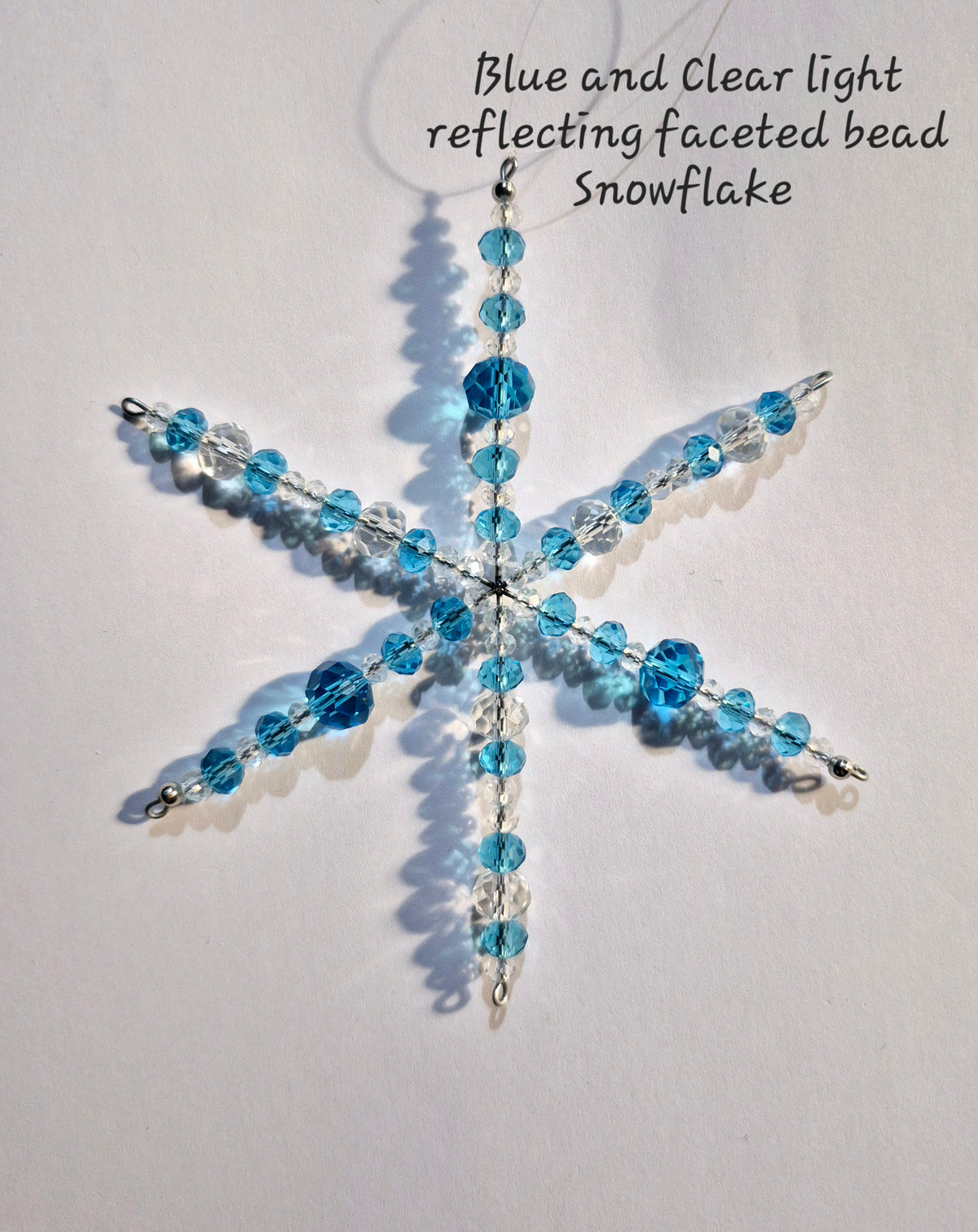 A handmade snowflake ornament made of faceted blue and clear beads on a silver wire frame, photographed against a plain white background with a text overlay explaining the bead properties.