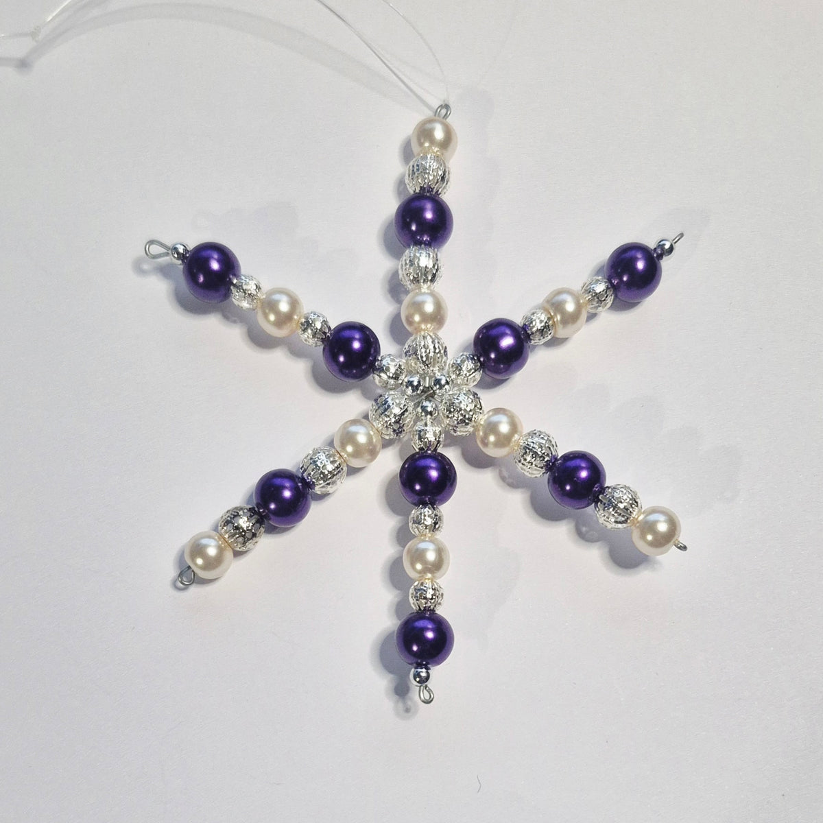 A beaded snowflake Christmas ornament featuring purple beads, white faux pearls, and silver rhinestone accents, suspended by a clear string on a light gray background.