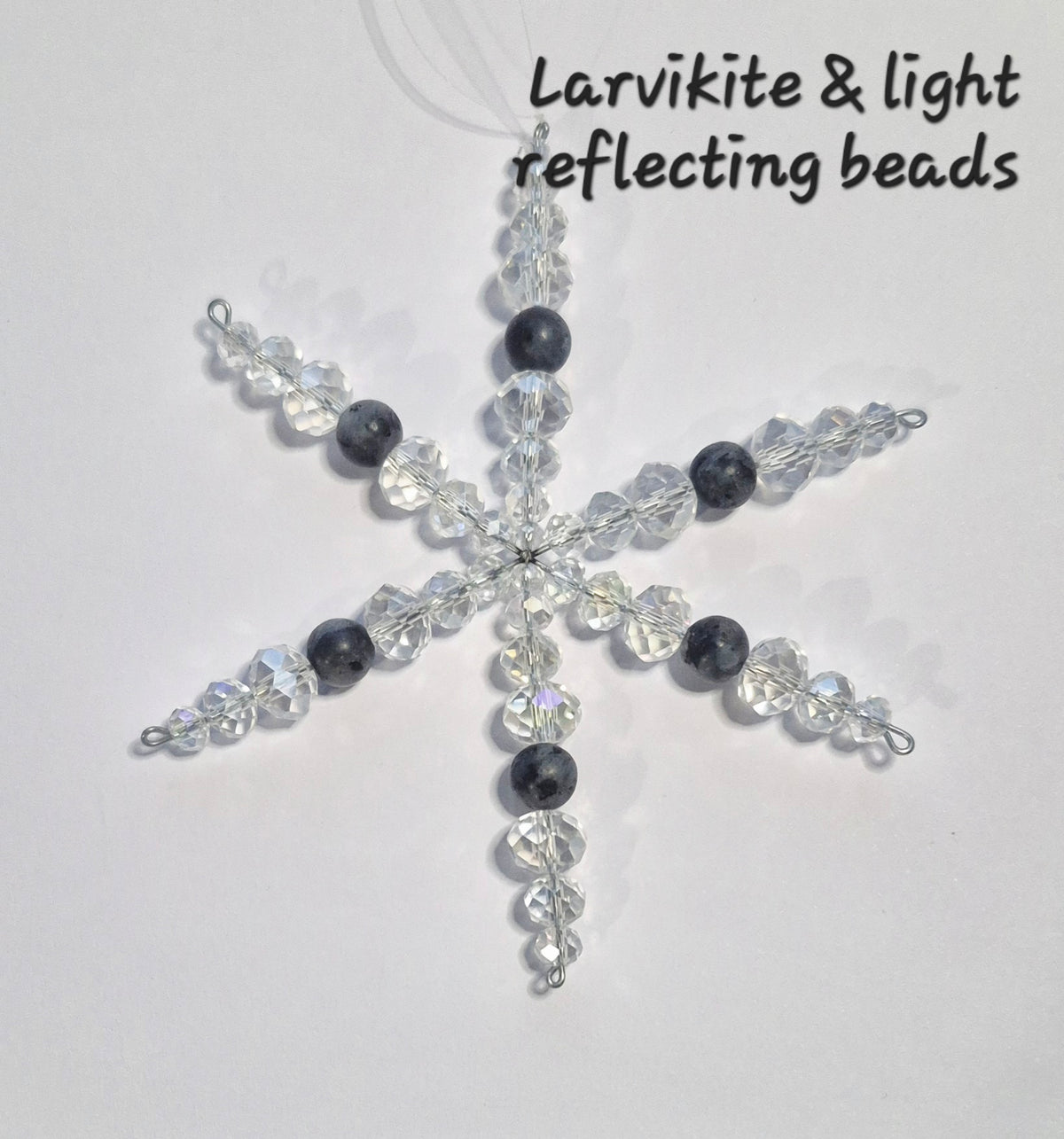 A snowflake-shaped beaded ornament made of clear faceted beads and dark gray round beads, with black text in the upper right reading 'Larvikite & light reflecting beads' on a plain light background.