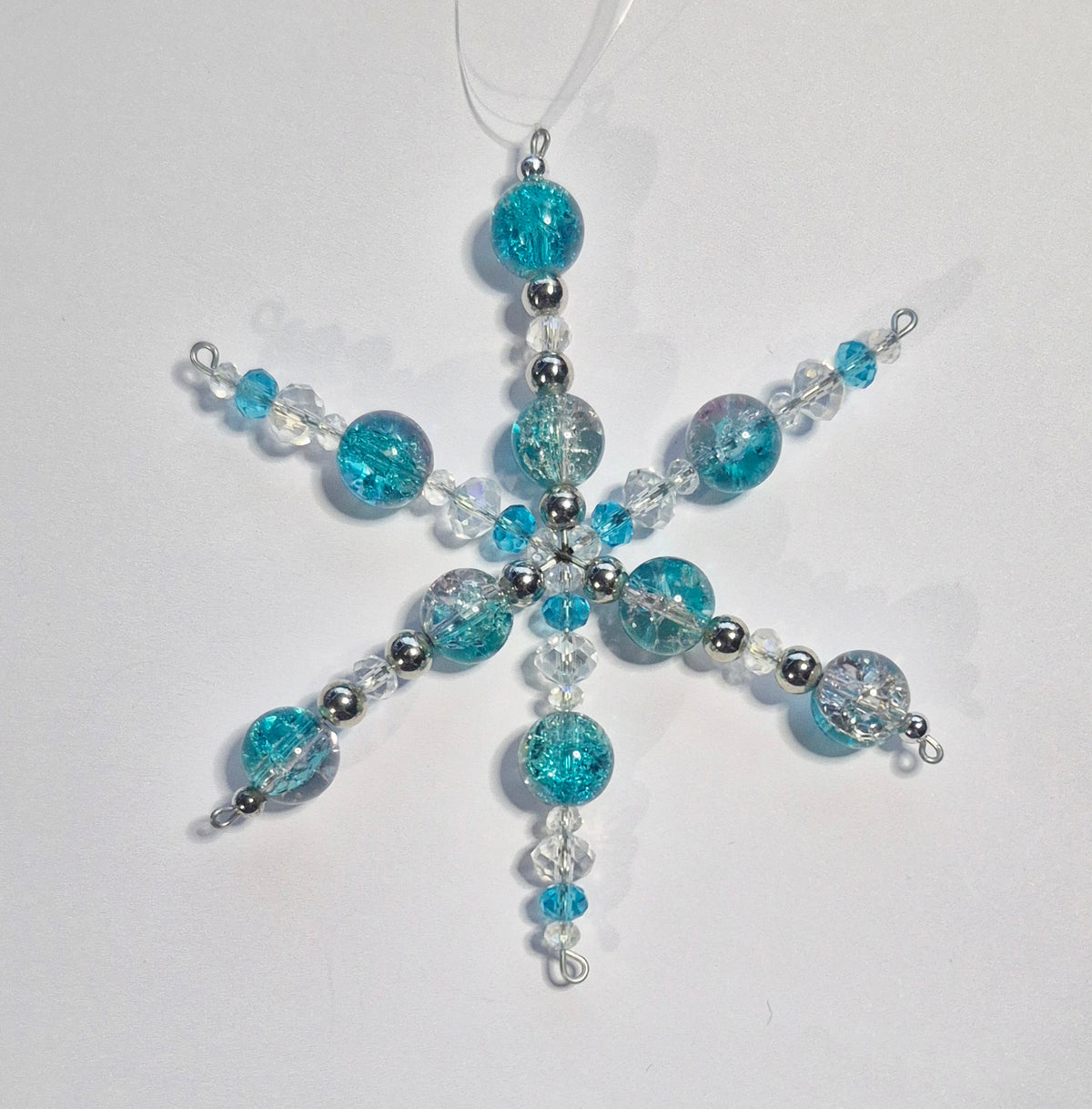 A handmade beaded snowflake ornament featuring turquoise blue, clear, and silver beads, suspended from a white string against a plain white background.