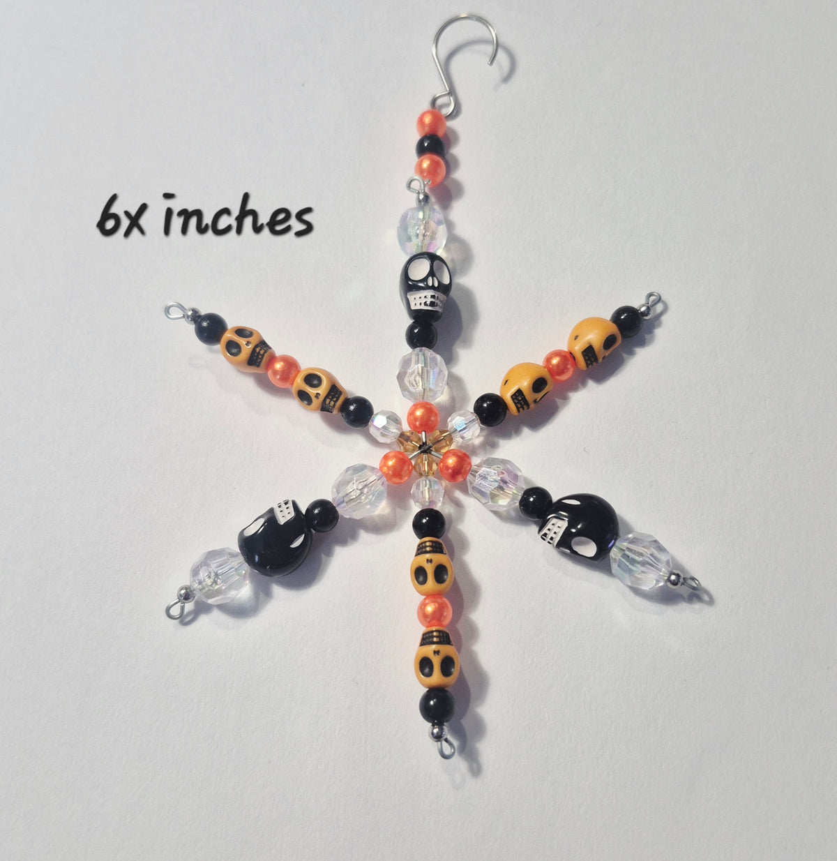 A handmade beaded snowflake ornament with Halloween-themed beads, including black skulls, orange and black patterned beads, and clear faceted beads, shown on a light gray background with '6x inches' text overlay.