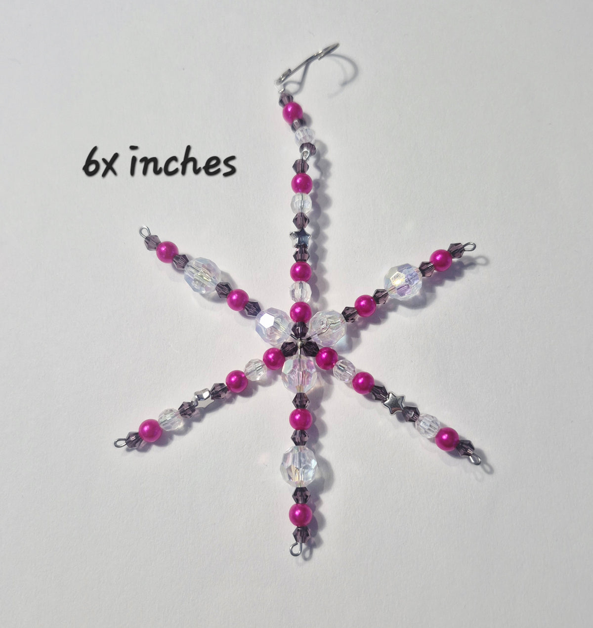 A handmade beaded snowflake ornament with pink, purple, and clear faceted beads, hanging from a silver fishhook. The text '6x inches' is written on the left side against a plain light background.