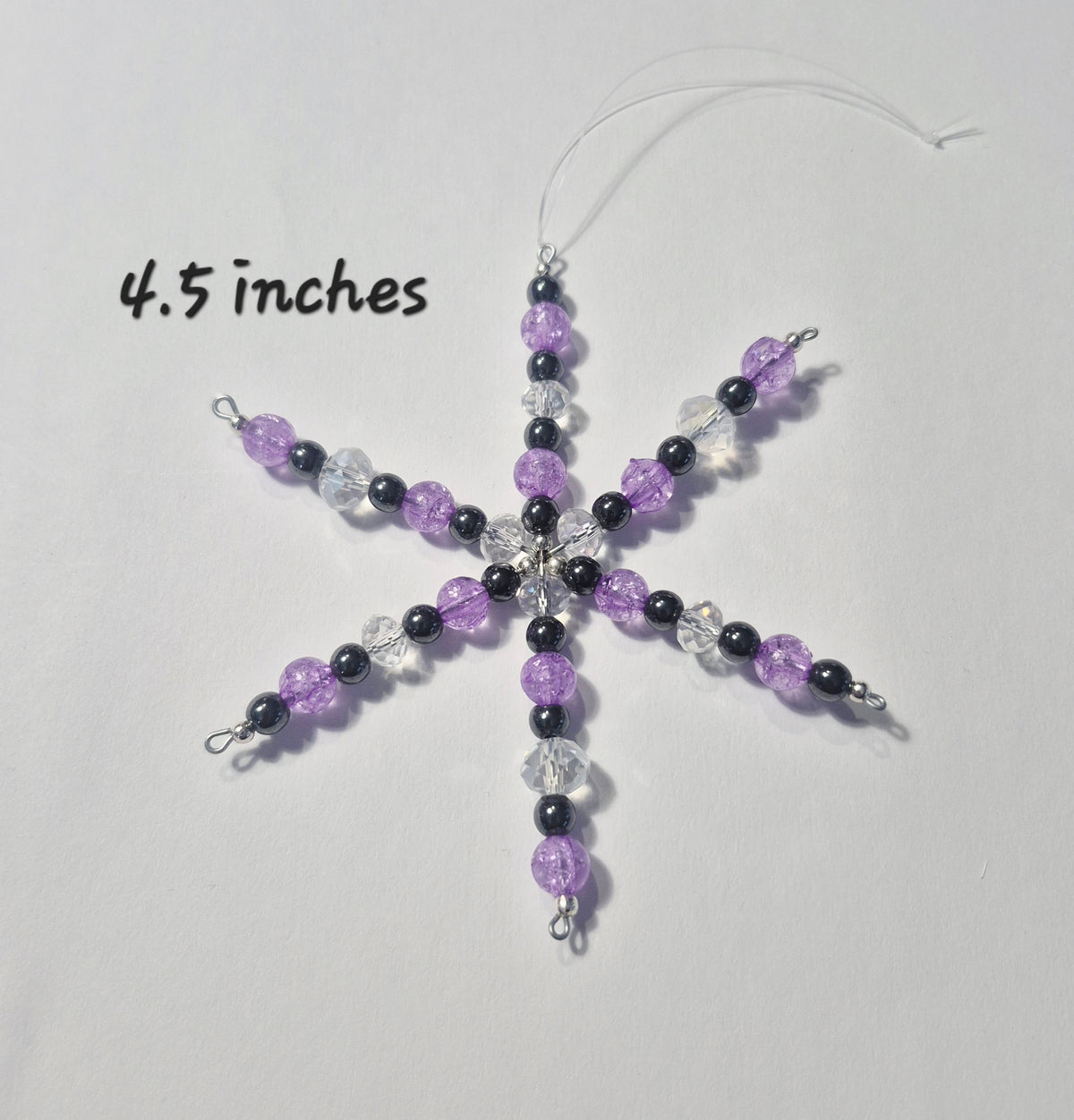 A handmade beaded snowflake ornament in purple, black, and clear beads, shown on a light gray background with a hanging string and a '4.5 inches' size text overlay.