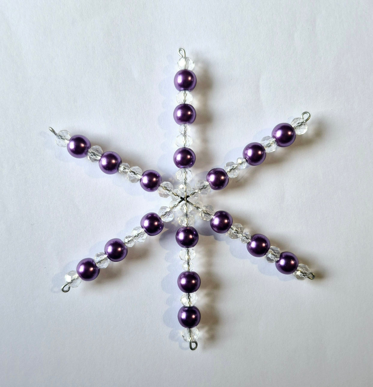 A six-pointed snowflake ornament made of purple faux-pearl beads and clear faceted beads on a silver wire frame, displayed on a plain white background.