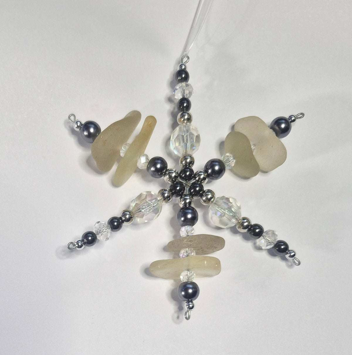 A handmade snowflake ornament featuring sea glass beads, dark gray pearls, and clear faceted beads on a white background.