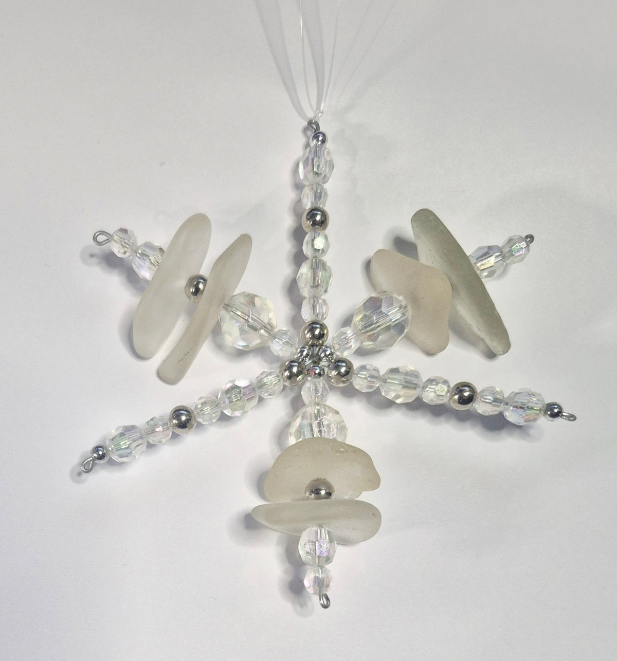 A handmade snowflake ornament made from clear faceted beads, silver beads, and semi-opaque white sea glass pieces, hanging from a string of clear line on a plain white background.