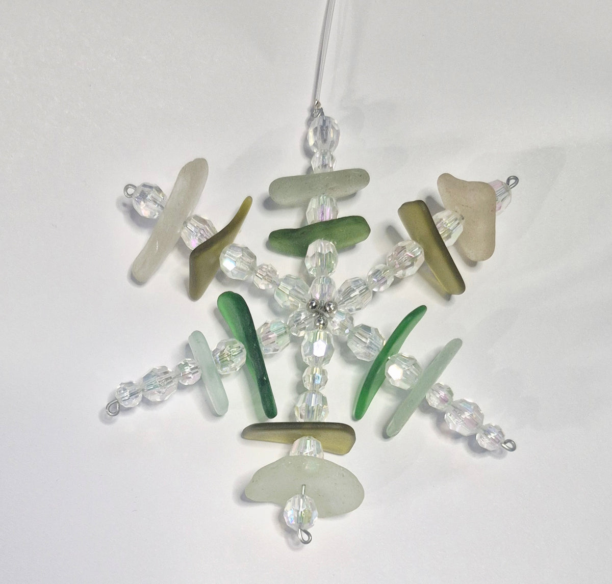 A handmade snowflake ornament made from clear faceted beads and natural sea glass pieces in shades of green, beige, and gray, hanging from a string of clear line against a plain white background.