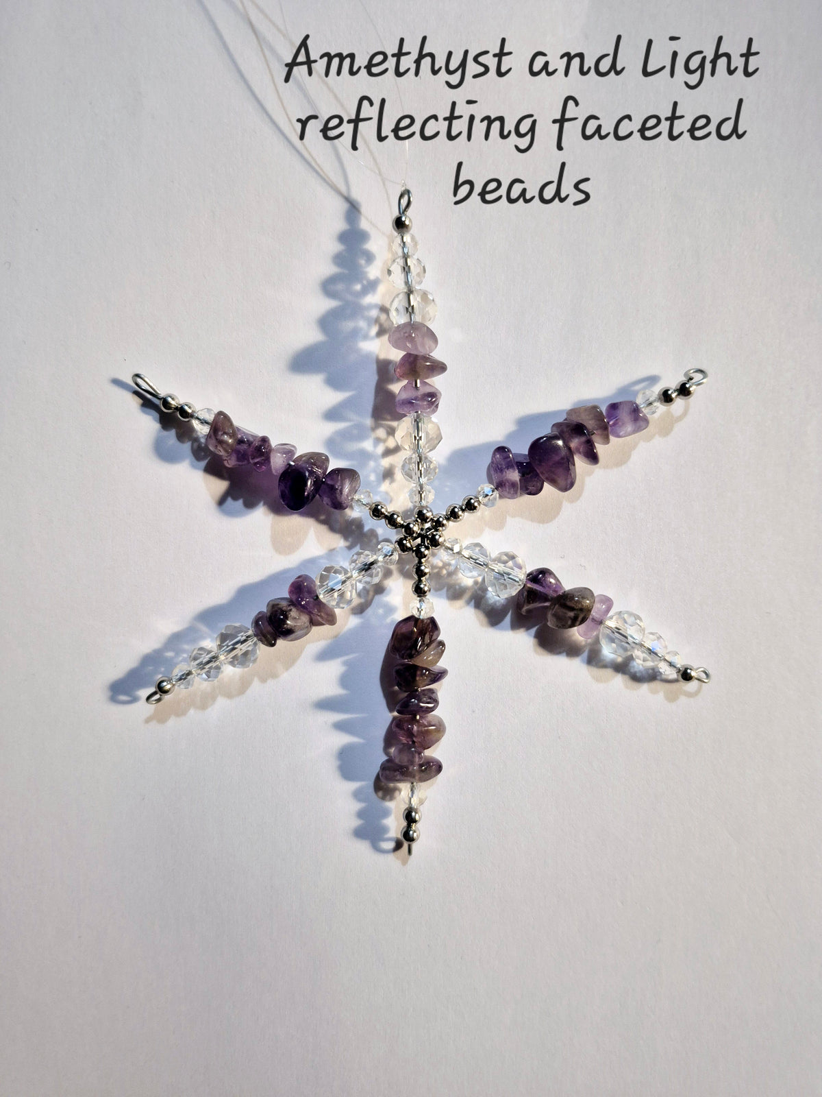 A snowflake-shaped hanging ornament made from purple amethyst chips, clear faceted beads, and silver-toned metal, photographed on a plain white background with handwritten text at the top reading 'Amethyst and Light reflecting faceted beads'.