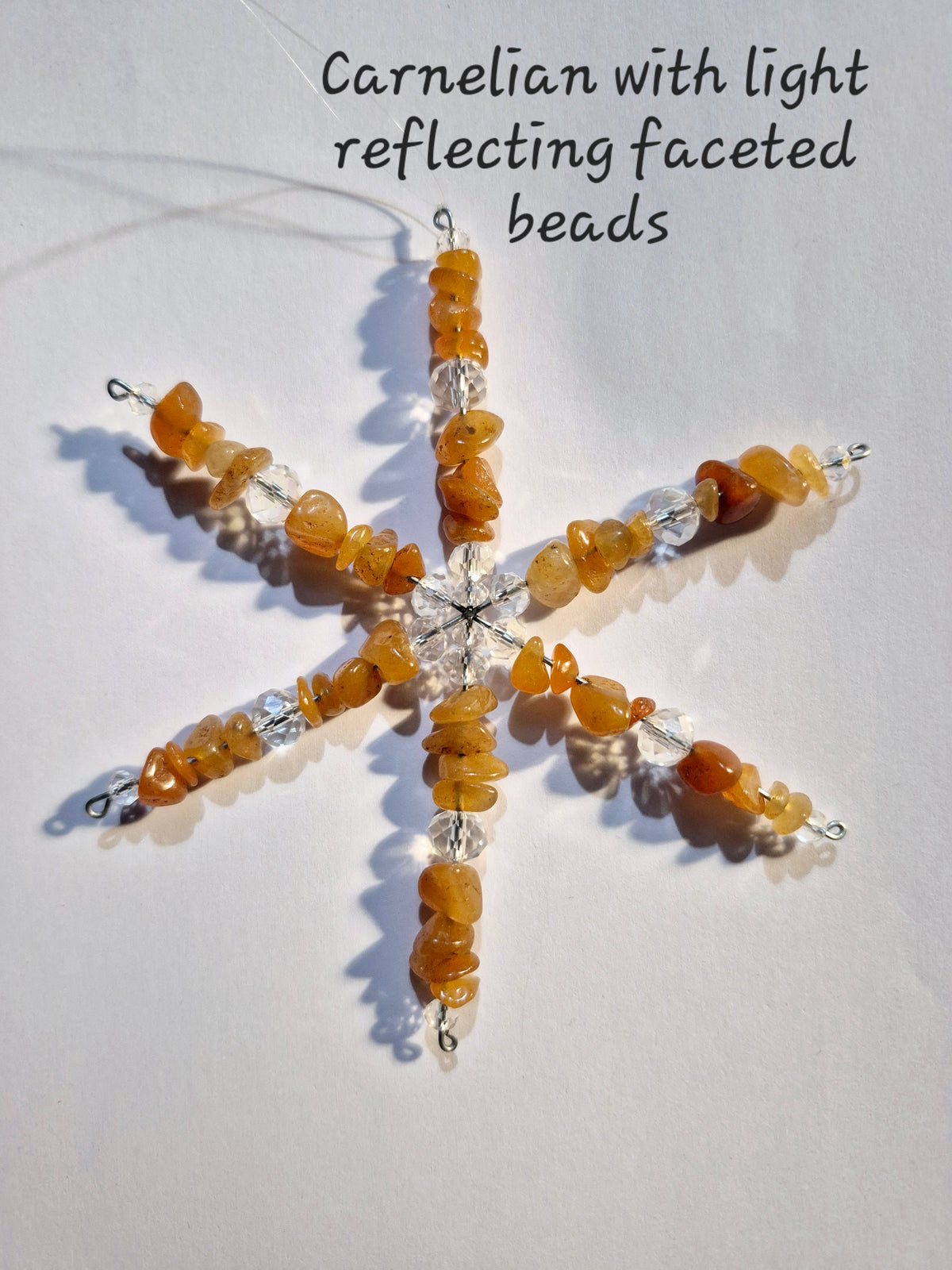 A star-shaped beaded ornament made of warm amber-orange faceted carnelian beads and clear reflective beads, hanging against a plain white background with text reading 'Carnelian with light reflecting faceted beads.'.