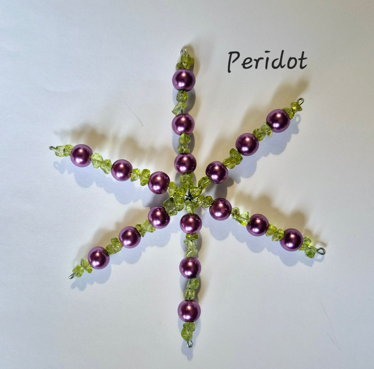 A handmade beaded snowflake ornament featuring metallic purple peridot beads and light green faceted beads, with the word 'Peridot' written in black text above it on a white background.