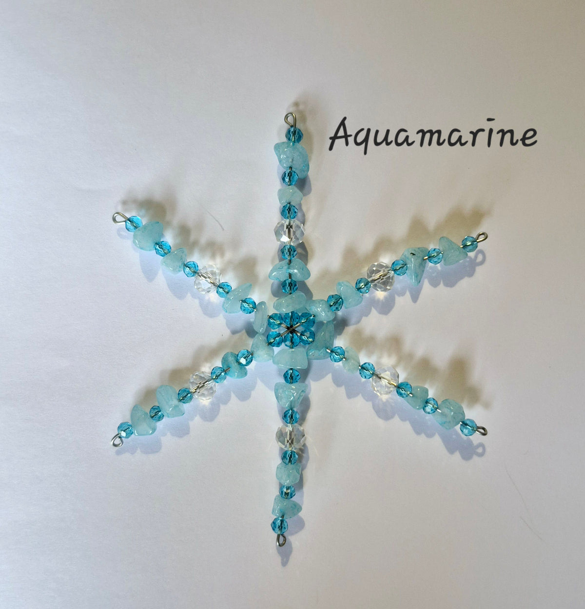 A handmade beaded snowflake ornament in aquamarine blue beads and clear faceted crystals, displayed on a plain off-white background with the text 'Aquamarine' written in black script.