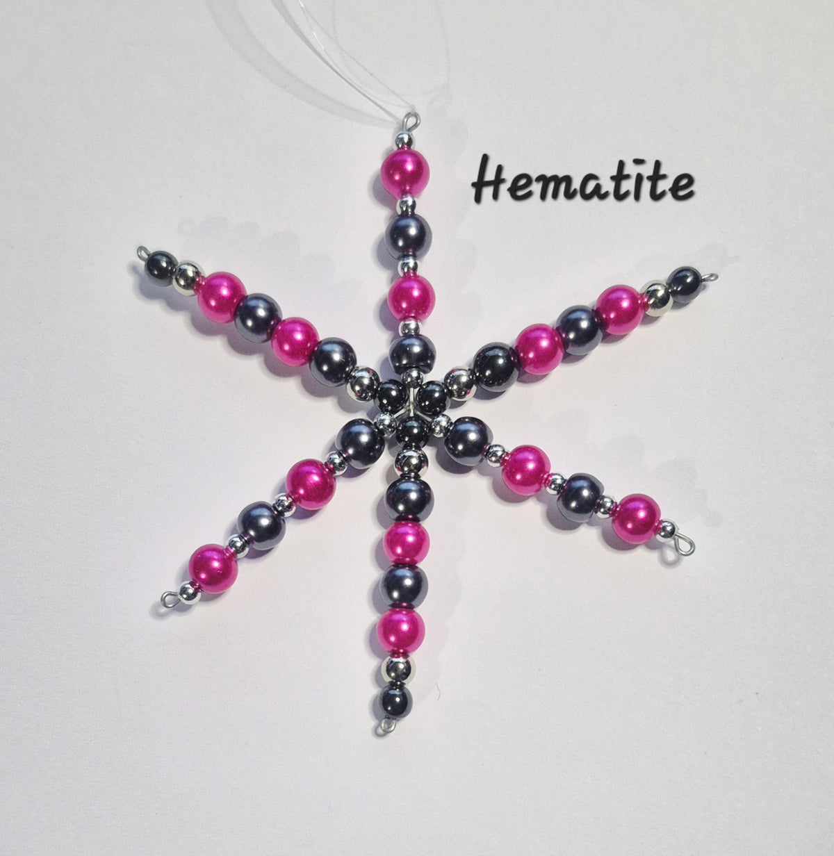 A handmade beaded snowflake ornament made of pink, dark gray hematite, and silver beads, hanging from a clear string. The word 'Hematite' is printed in black text on the right side of the image.