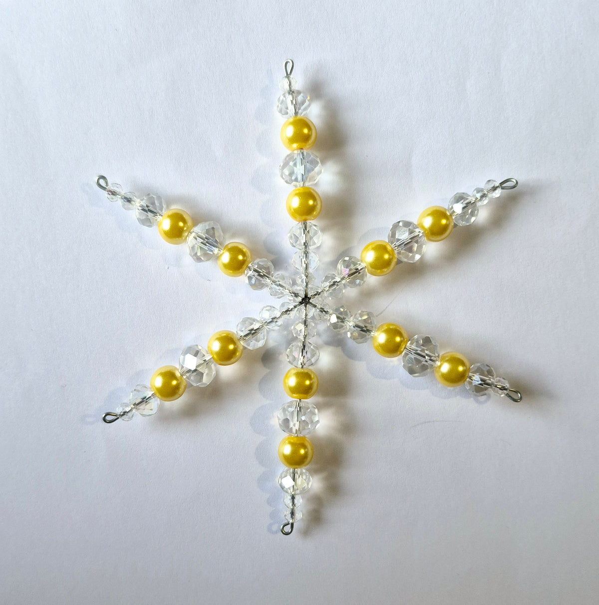 A six-pointed snowflake ornament made of yellow faux-pearl beads and clear faceted beads on a silver wire frame, displayed on a plain white background.