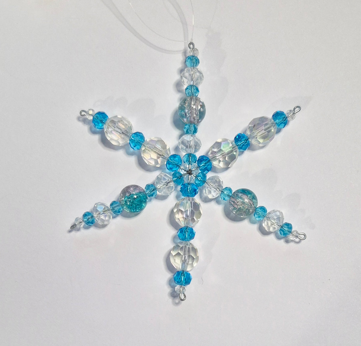 A handmade beaded snowflake ornament featuring light blue and clear faceted beads on a white background.
