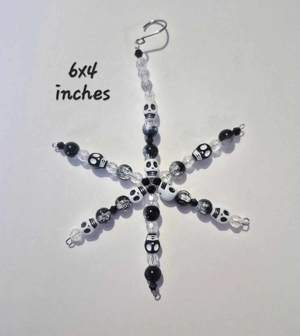 A handmade beaded snowflake ornament featuring black, white, and clear skull beads, with a silver fishhook clasp at the top. Handwritten text in the upper left indicates a size of 6x4 inches.