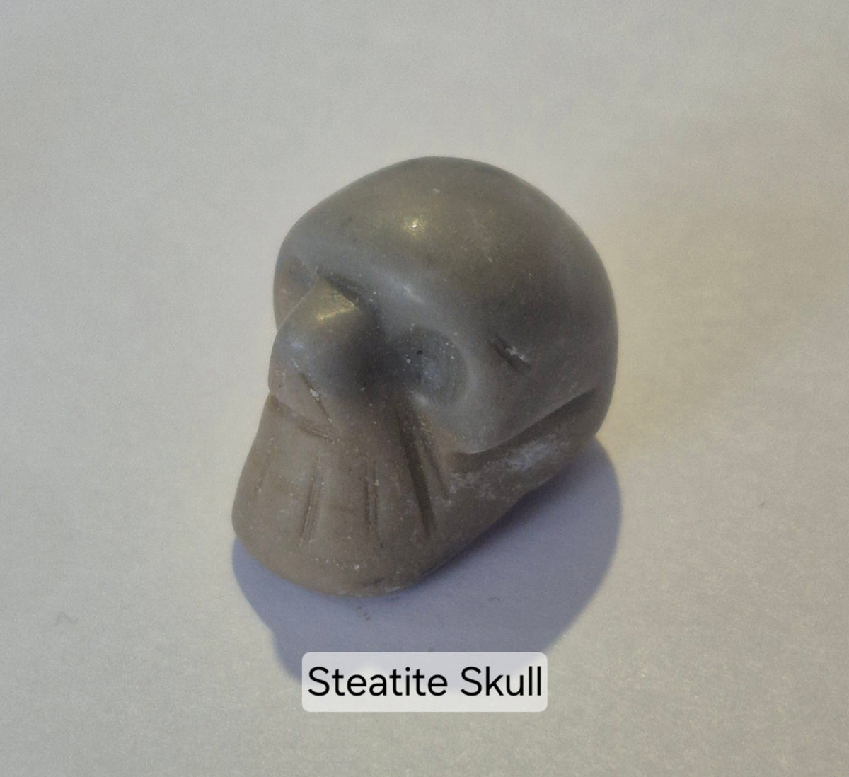A steatite skull figurine with a smooth matte gray finish, shown on a light gray background with a text label 'Steatite Skull' at the bottom.