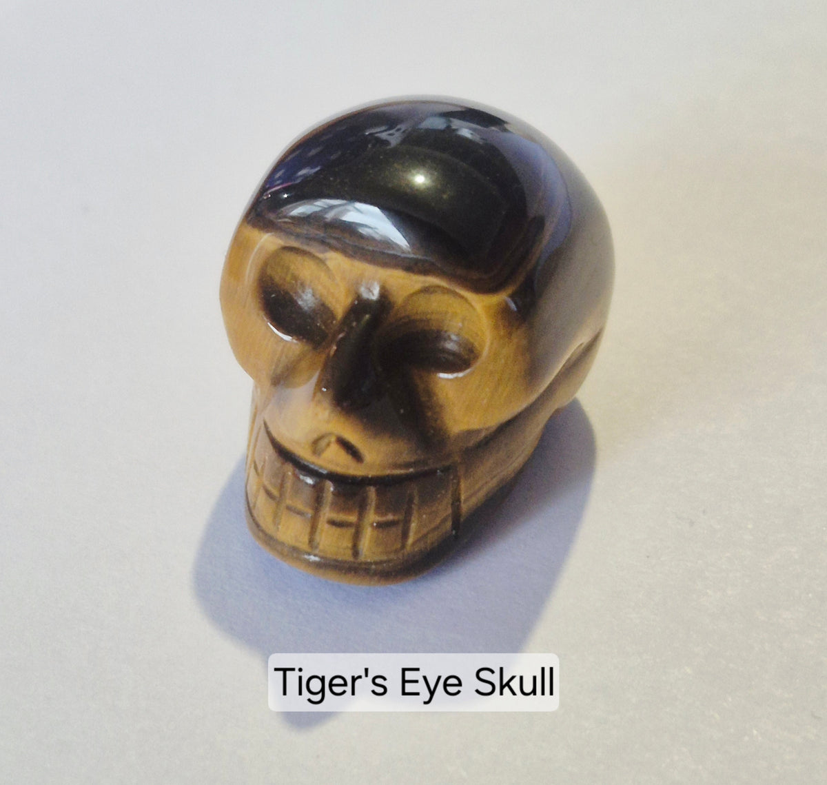 A carved tiger's eye skull bead or charm, showcasing its characteristic brown and black banded pattern, set against a plain light background with a text overlay reading 'Tiger's Eye Skull'.