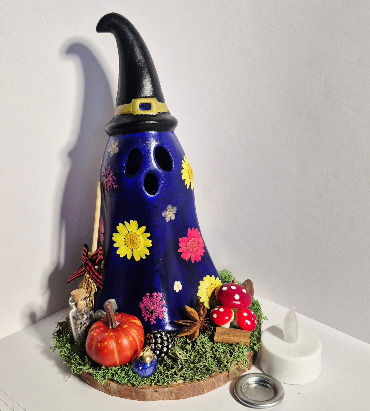 A handmade decorative Halloween ghost burner with a dark blue body, black pointed hat, and colourful floral and autumn accents, displayed on a white surface against a light gray background.