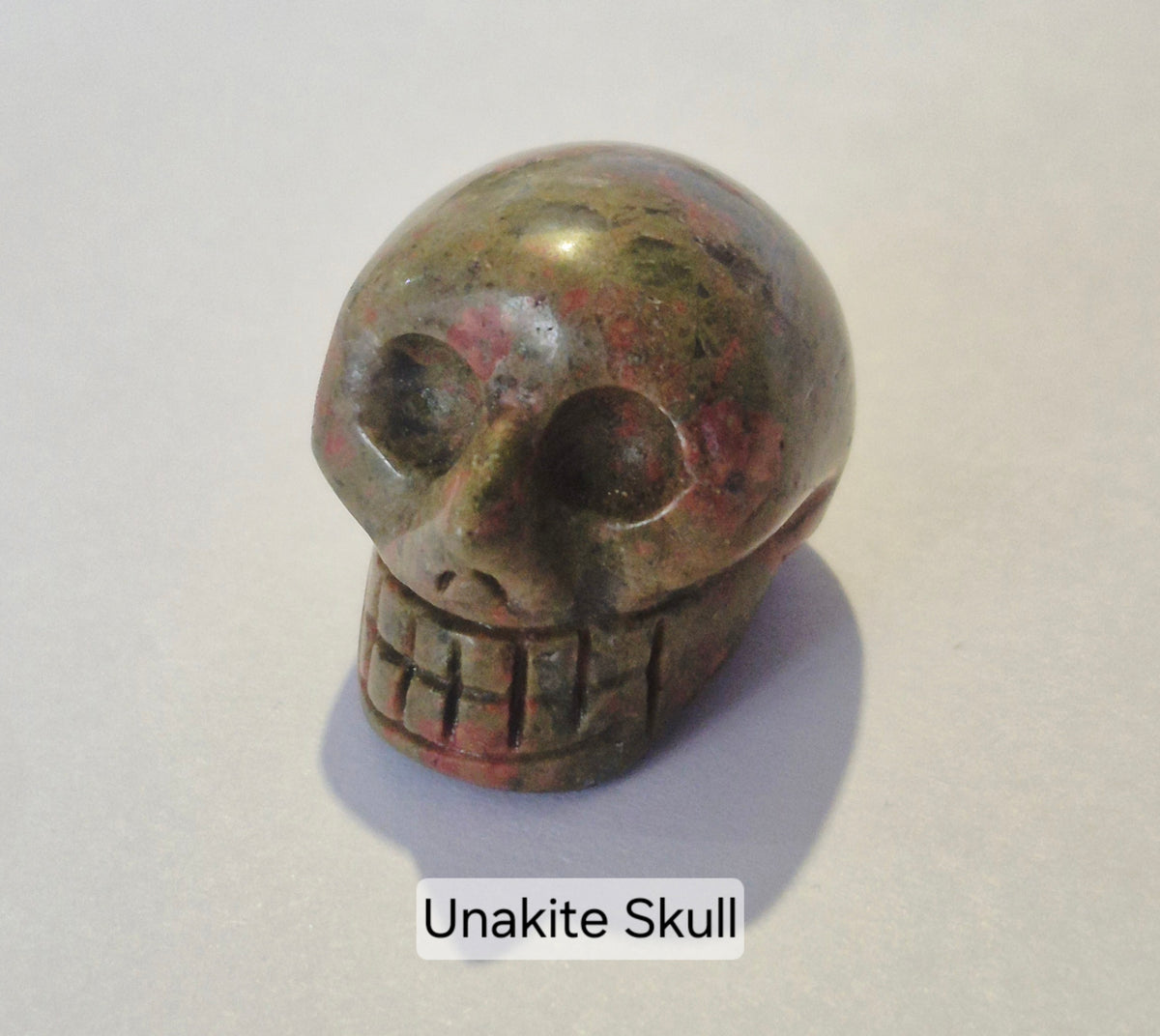 A carved unakite skull figurine with a polished, rounded surface showing natural earthy brown, green, and pinkish-red mineral variations. The skull is positioned on a light gray background with a soft shadow, and a text label at the bottom reads 'Unakite Skull'.