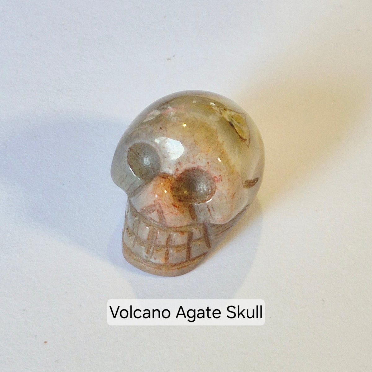 A small carved Volcano Agate skull figurine with natural earthy tones, shown on a plain light background with a text label below reading 'Volcano Agate Skull'.