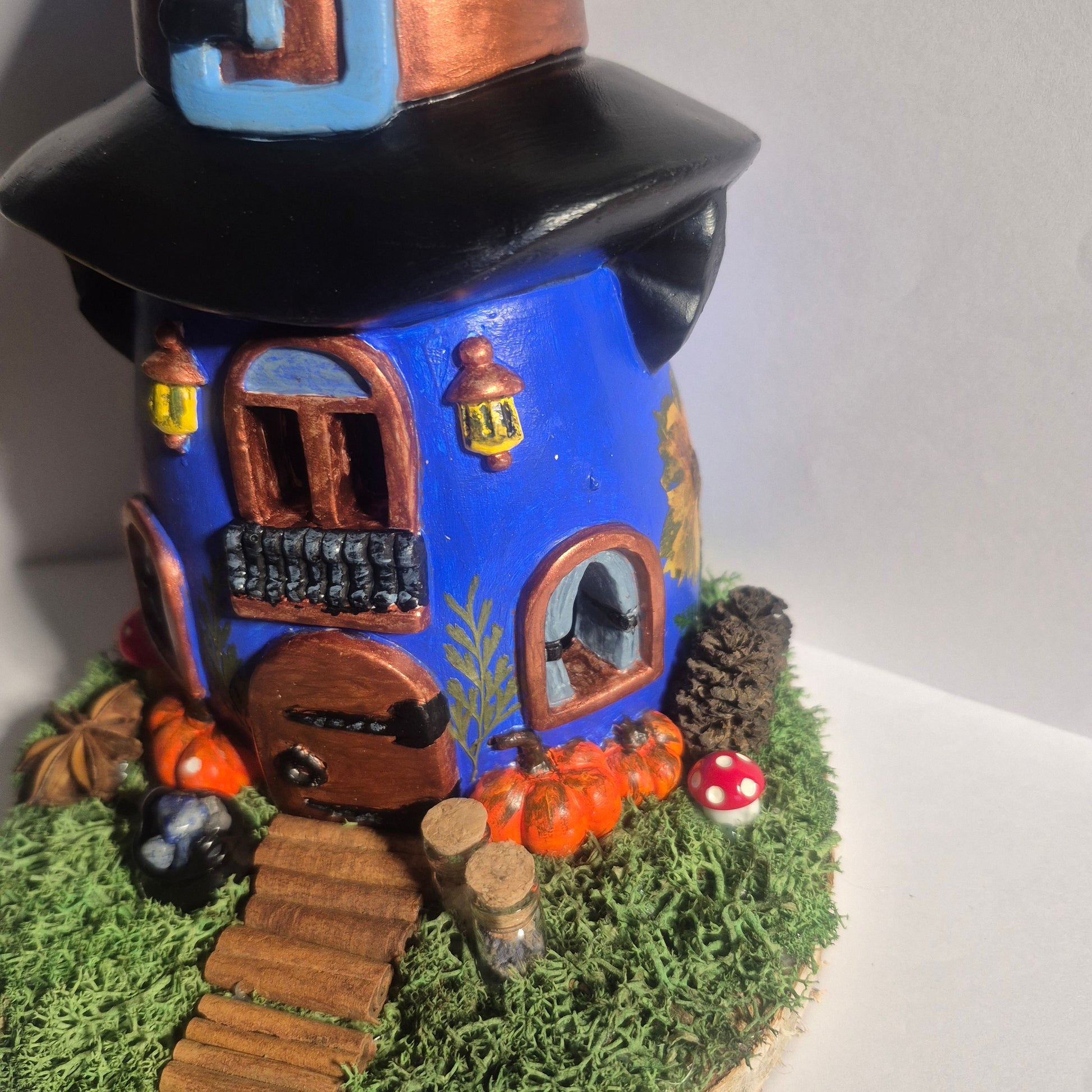 A handmade witch cottage figurine, with LED lights. The cottage has a dark blue body, a black pointed witch hat with a blue buckle, a brown door, and is decorated with faux moss, pumpkins, mushrooms, a path made of cinnamon, jars of lavender and celtic salt, and other magical details on a round wooden base.