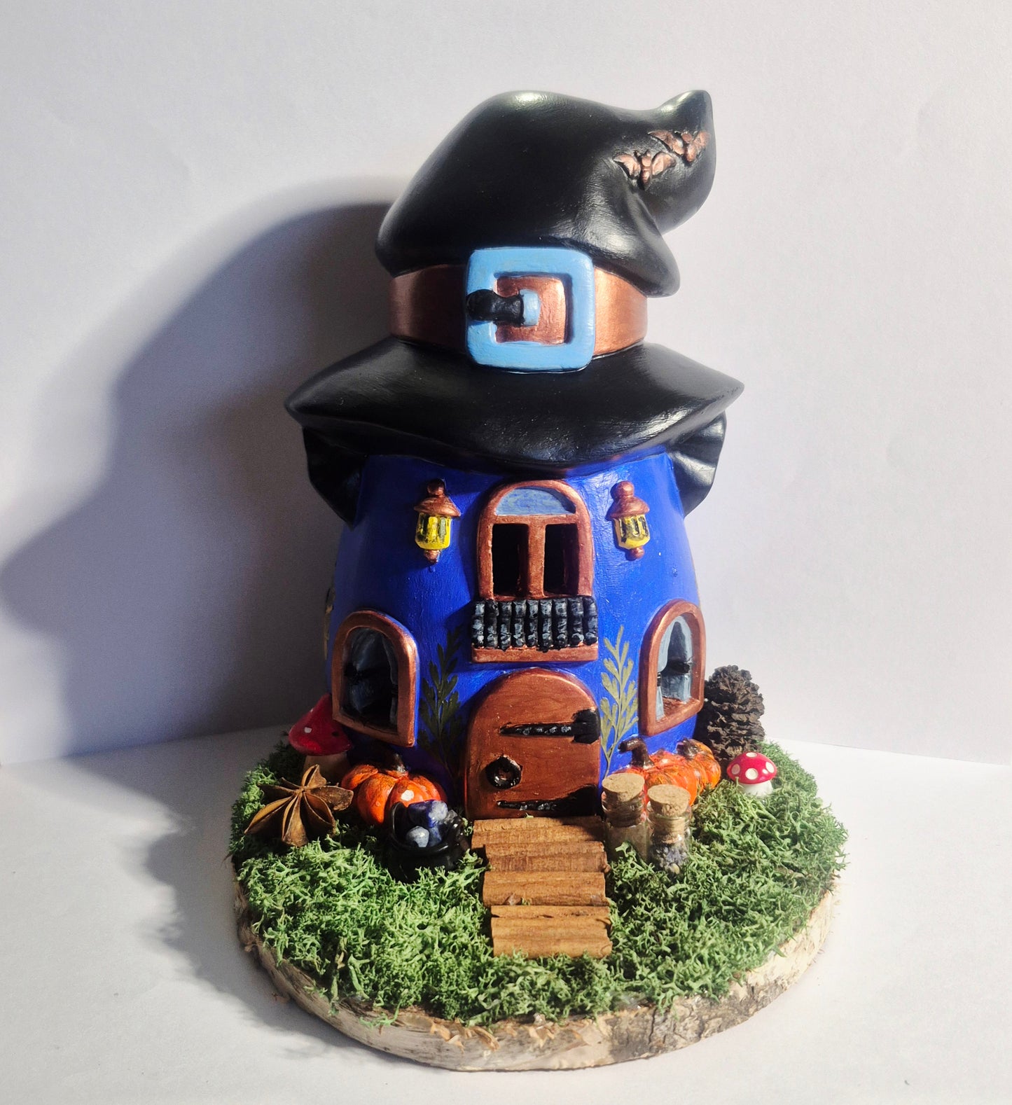 handmade and Hand-painted ceramic witch's cottage, with an LED candle inside, featuring a blue body, black hat, brown door, green moss base, real pressed flowers and magical decorations. 