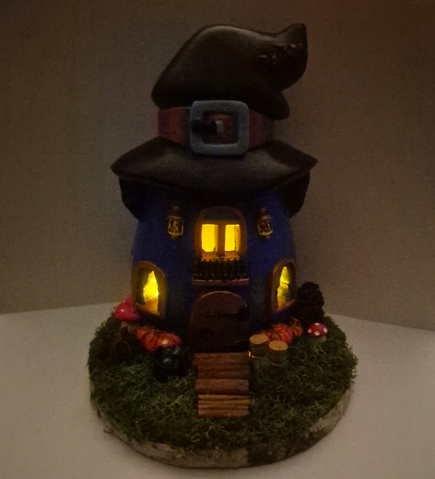 A handmade witch house figurine with a tall black hat, glowing yellow windows, a blue body, mossy green base, and magical decorative details, photographed against a plain indoor background.