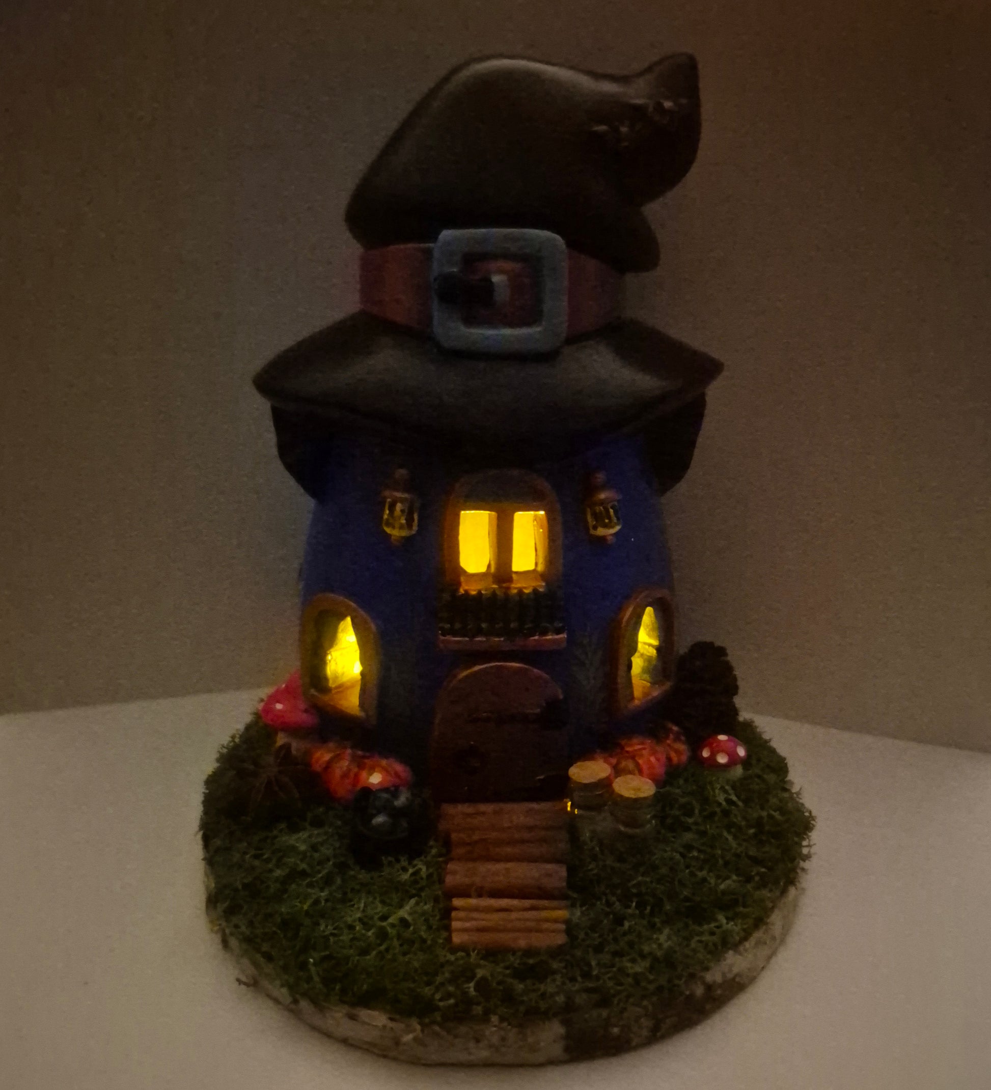 A handmade witch house figurine with a tall black hat, glowing yellow windows, a blue body, mossy green base, and magical decorative details, photographed against a plain indoor background.