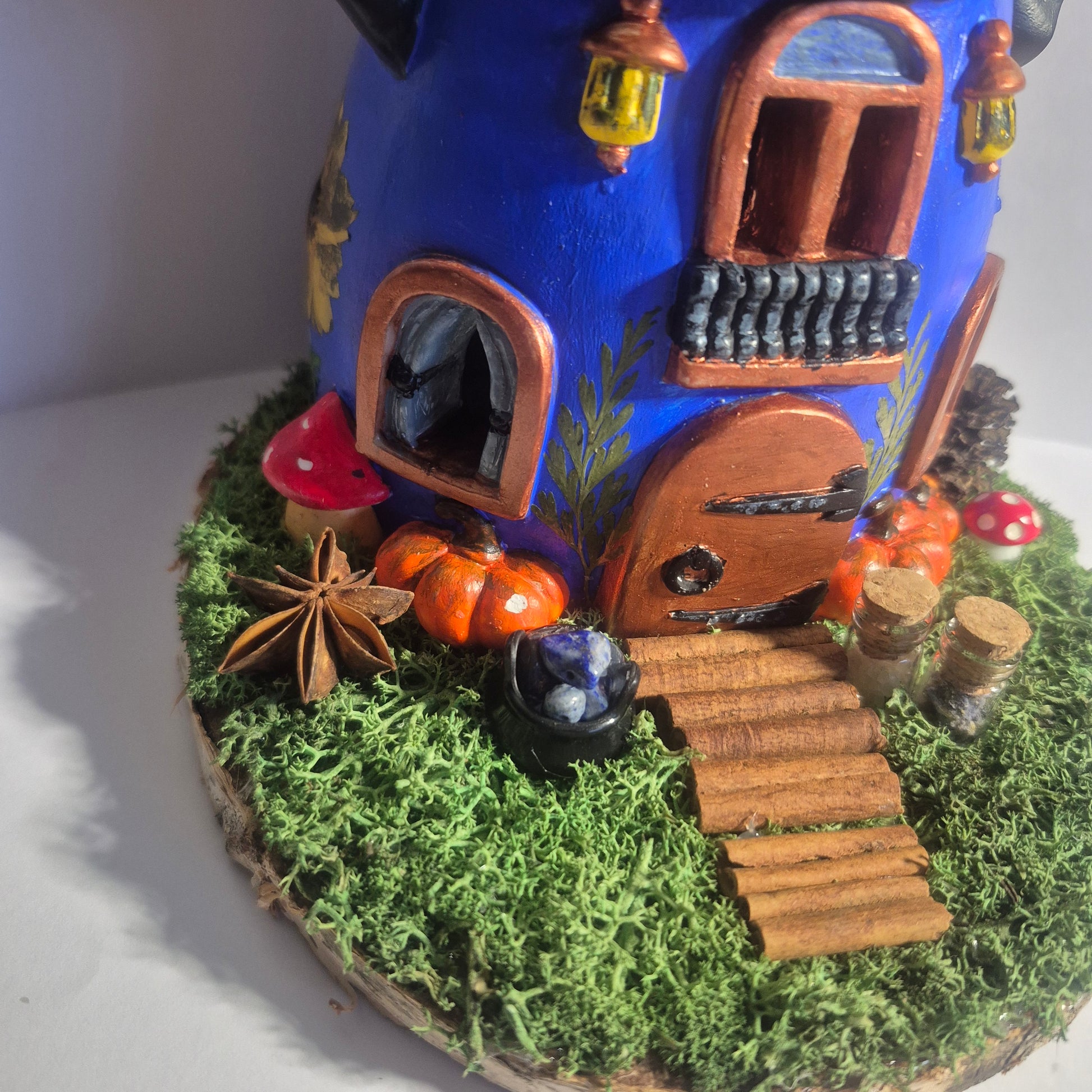 A handmade witch cottage figurine, with LED lights. The cottage has a dark blue body, a black pointed witch hat with a blue buckle, a brown door, and is decorated with faux moss, pumpkins, mushrooms and other magical details on a round wooden base.