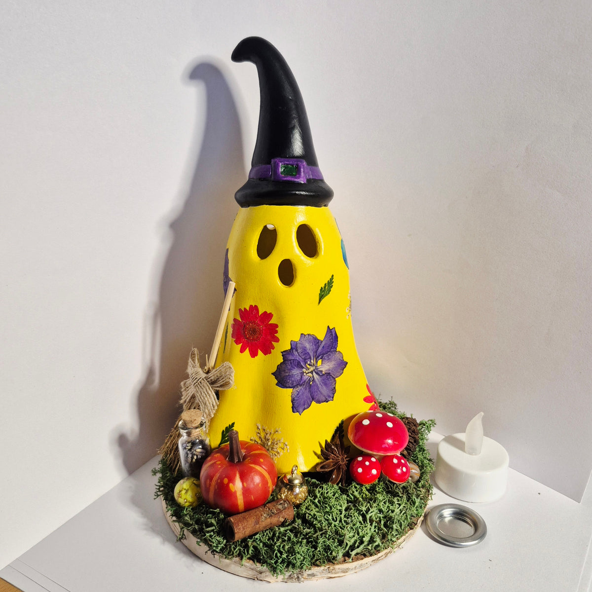 A Handmade decorative ghost-shaped candle holder with a black witch hat, yellow ghost body, and colorful floral and autumn accents, displayed on a white surface against a white wall.